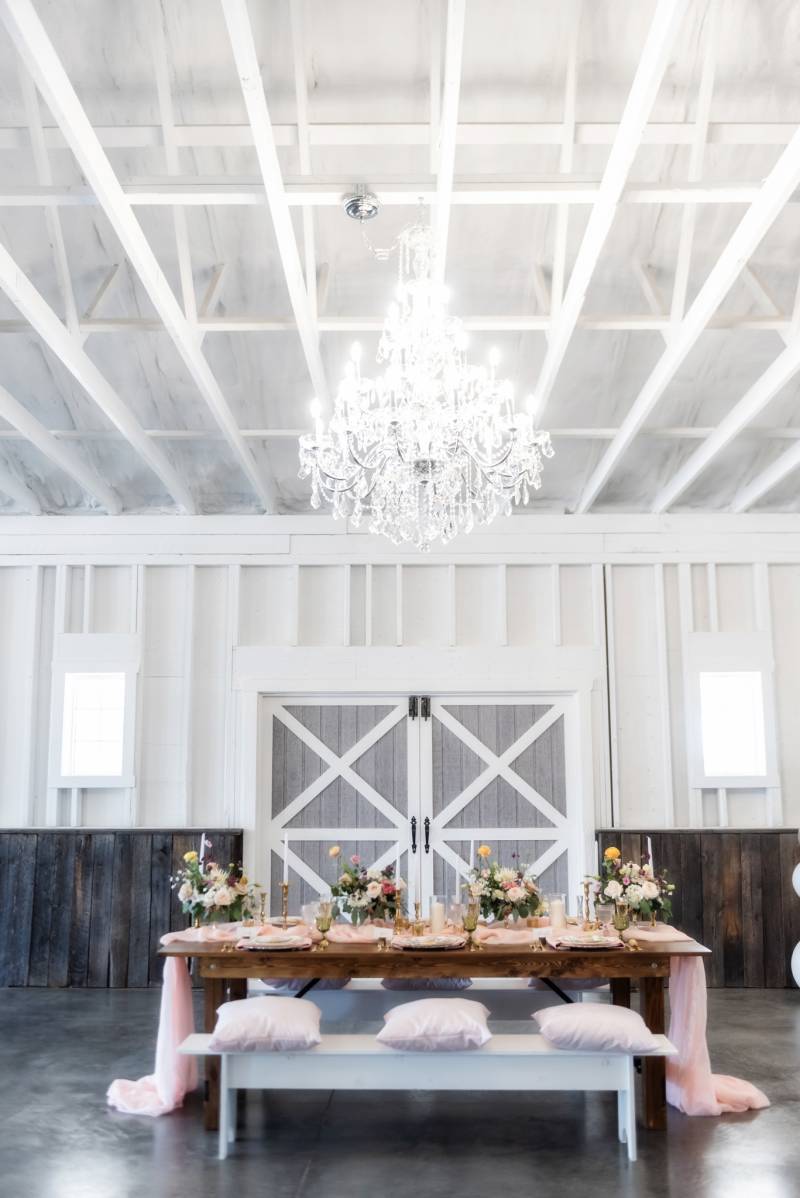 Large glass chandelier over wooden table with benches and blush table runner draping over side 