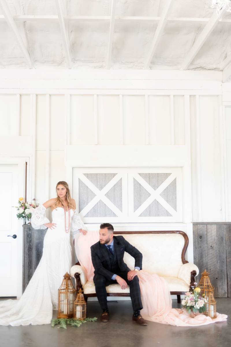 Groom sits on white sofa while bride stands leaning on back behind with draping pink fabric 