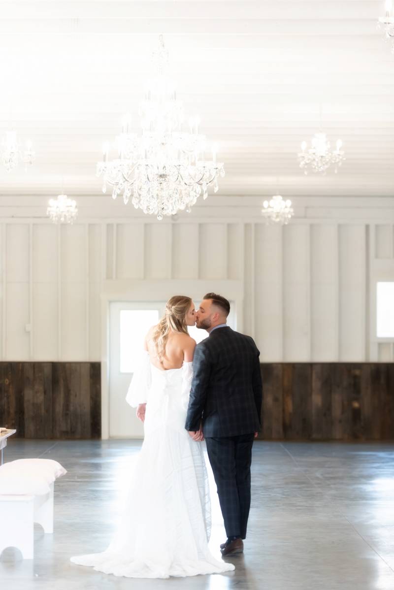 Bride and groom walk in large room holding hands kissing under glass chandelier 