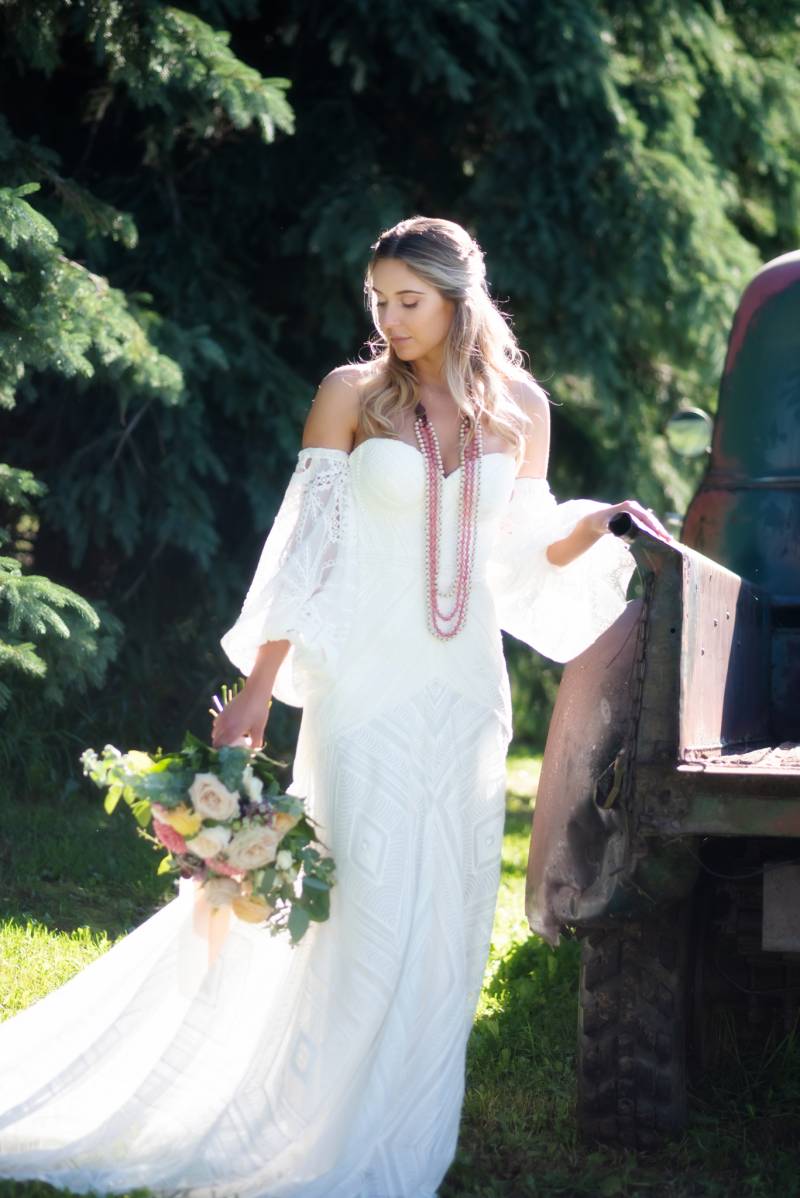 Bride standing holding onto truck bed in white shoulder less dress holding white and pink bouquet 