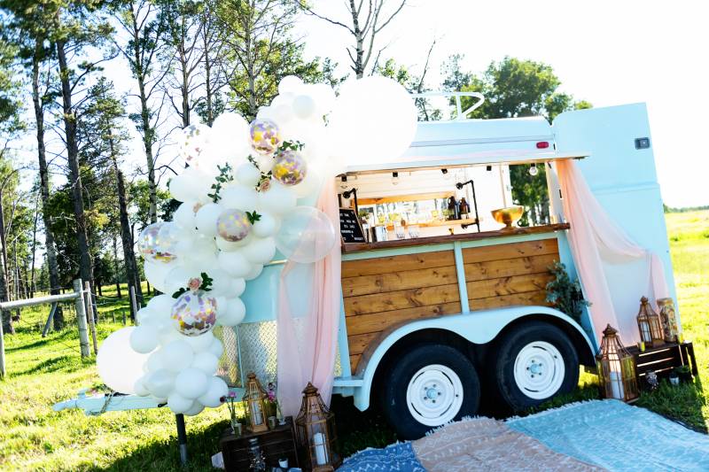 Mobile bar with white balloon arrangement and colored rugs below in grassy field 