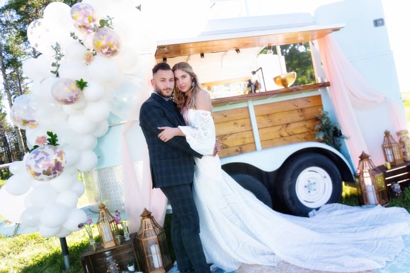 Bride and groom embrace in front of mobile bar with white balloon arrangement 