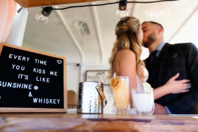 Bride and groom kiss behind bar table with black sign and drinks 