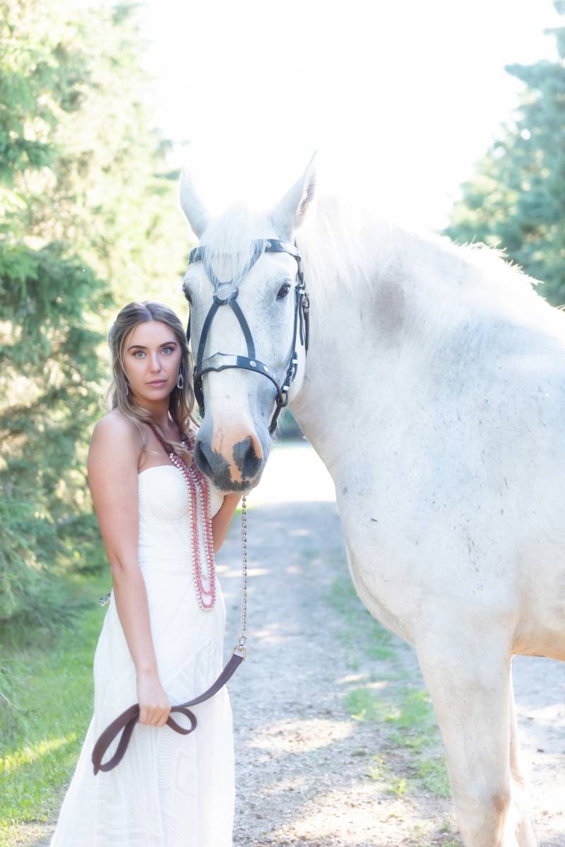 Bride standing in white dress beside large white horse on gravel path