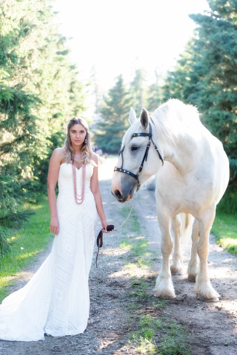 Bride standing inn white dress and hanging pink beads holding rope with white horse 