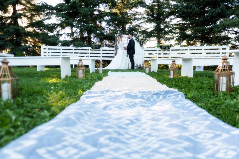 Bride and groom stand at the end of long white fabric laying on green grass backing white fence