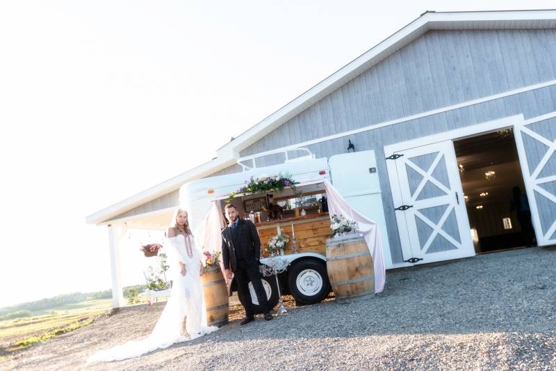 Mobile drink bar behind bride and groom standing in front of blue barn with open door 