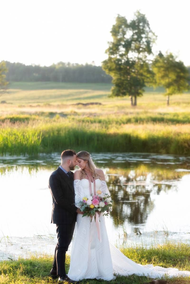 Bride and groom stand touching foreheads holding bouquet backing small pond
