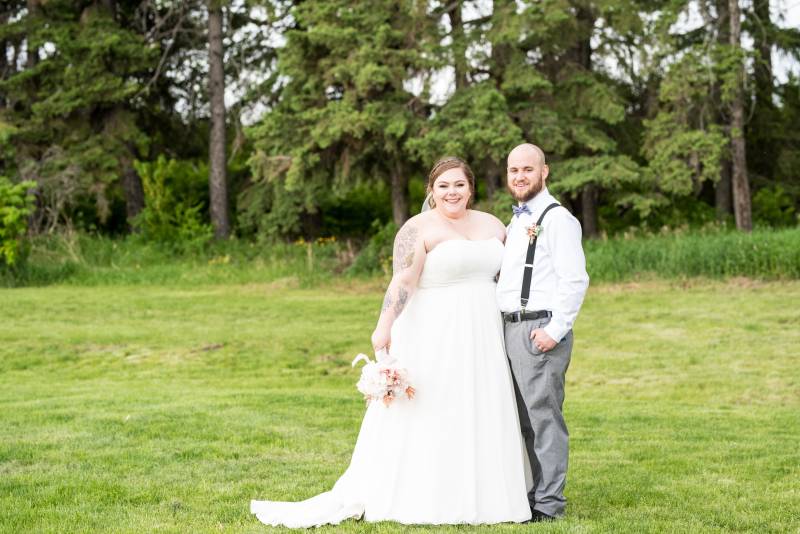 Bride and groom stand together in grassy field backing forest