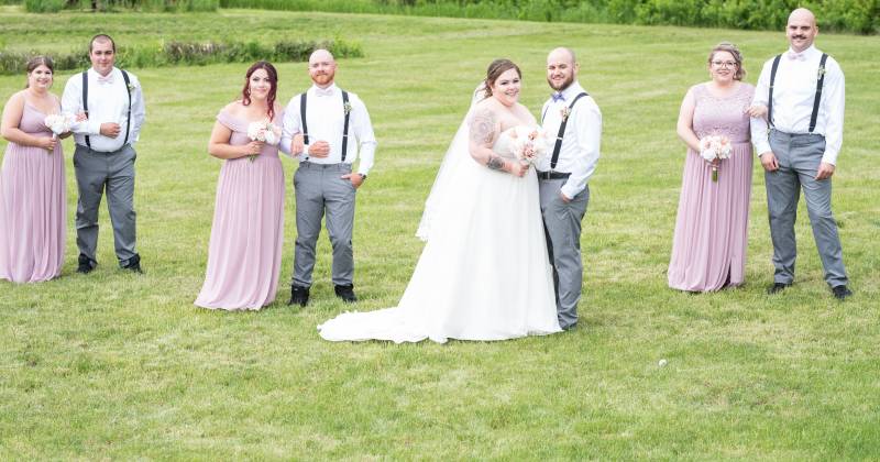 Bridesmaids and groomsmen stand in twos behind bride and groom in grassy field 