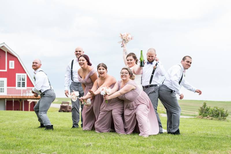 Bridesmaids and groomsmen and bride and groom laughing arms out in grassy field 