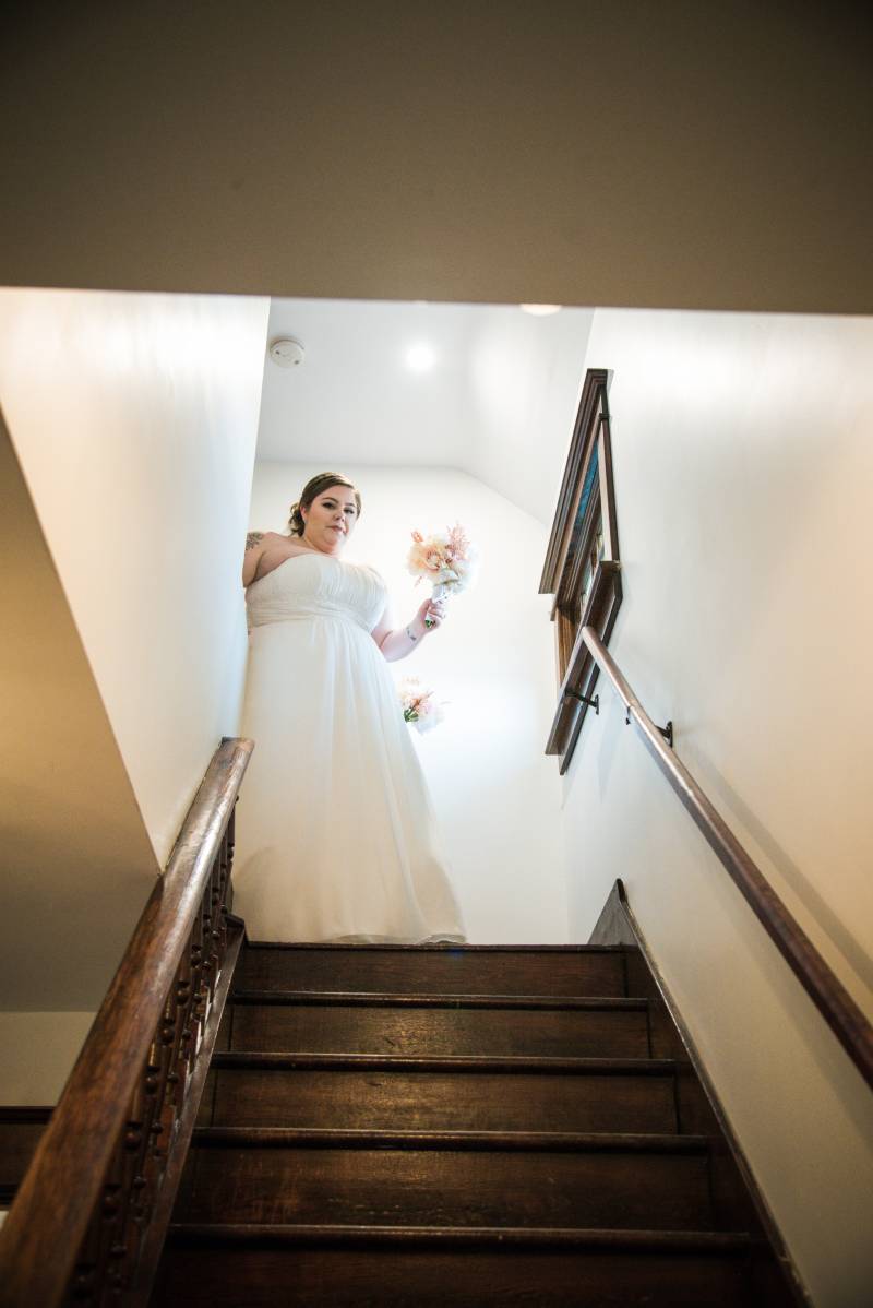 Woman in white dress holding pink and white bouquet walking down wooden stairwell 