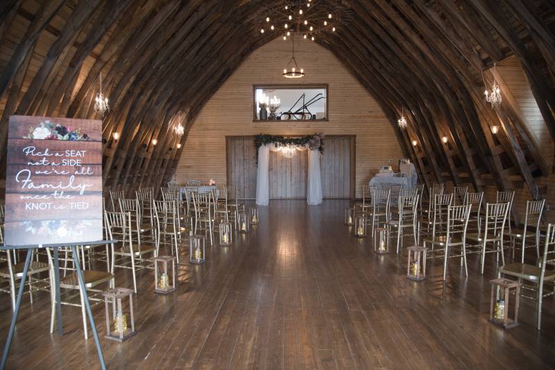 Inside large wooden barn with gold seats and wedding arch with hanging white fabric 
