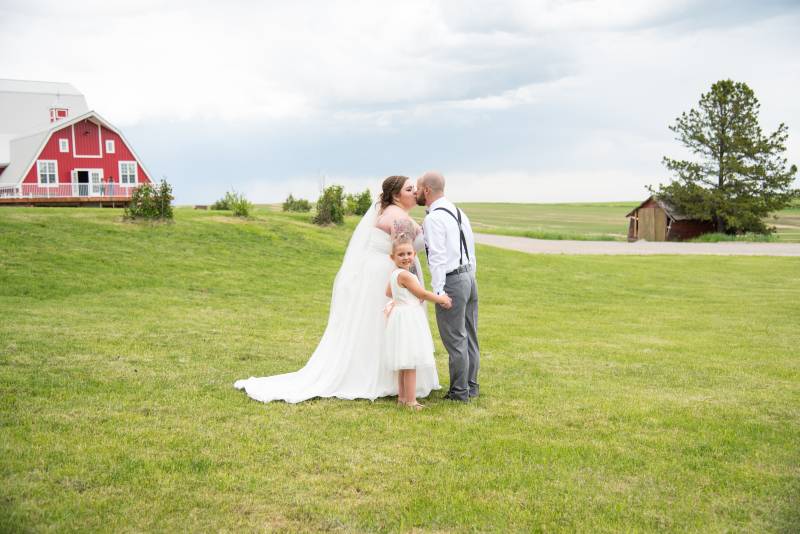Bride and groom kiss while flower girl stands holding hands in grassy field 