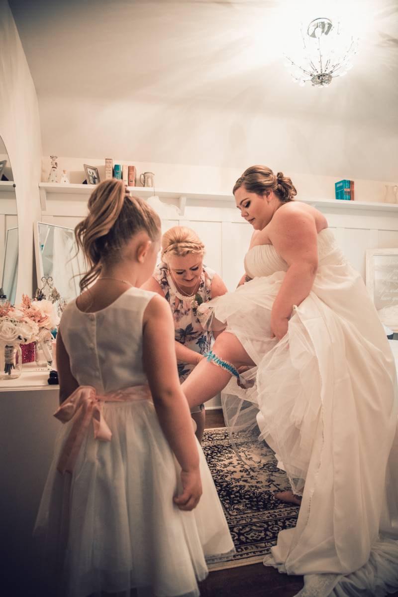 Woman in white dress has blue ribbon tied around calf as flower girl watches 