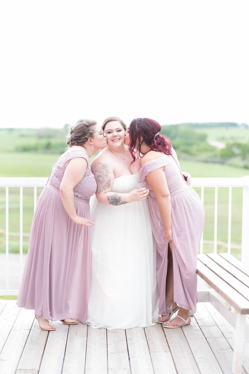 Bride being kissed by bridesmaids on either side standing on deck  