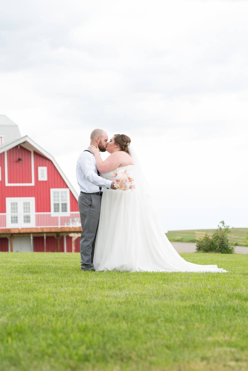 Bride and groom kiss in grassy field in front of red barn 
