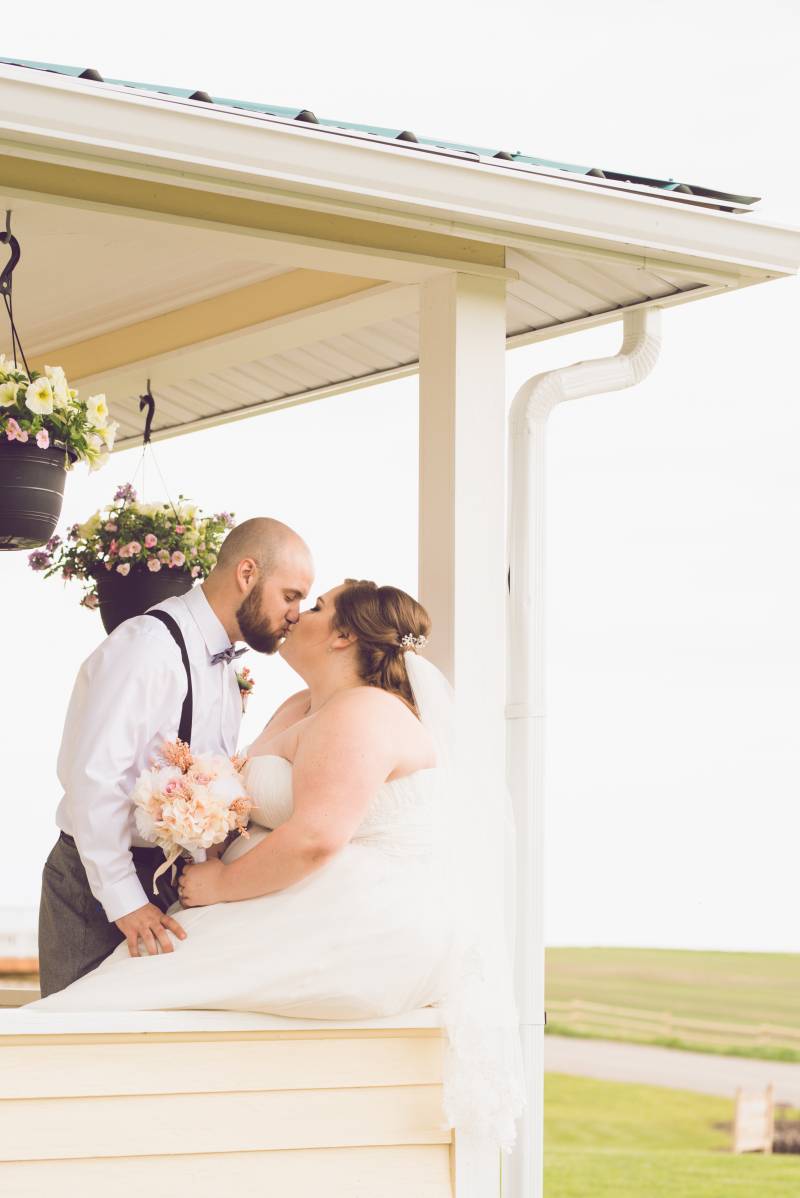 Bride and groom kiss sitting on ledge of  deck with hanging flowerpots 