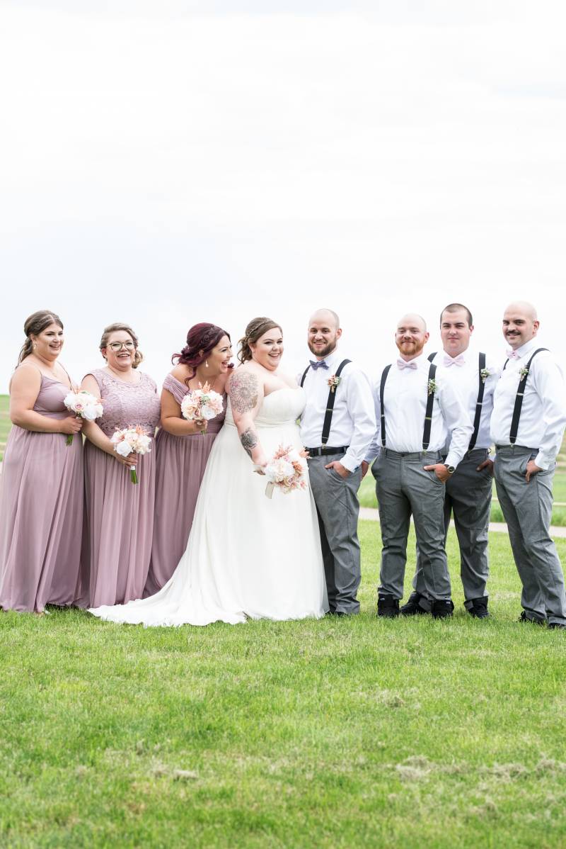 Bride and groom stand together beside bridesmaids and groomsmen in grassy field 