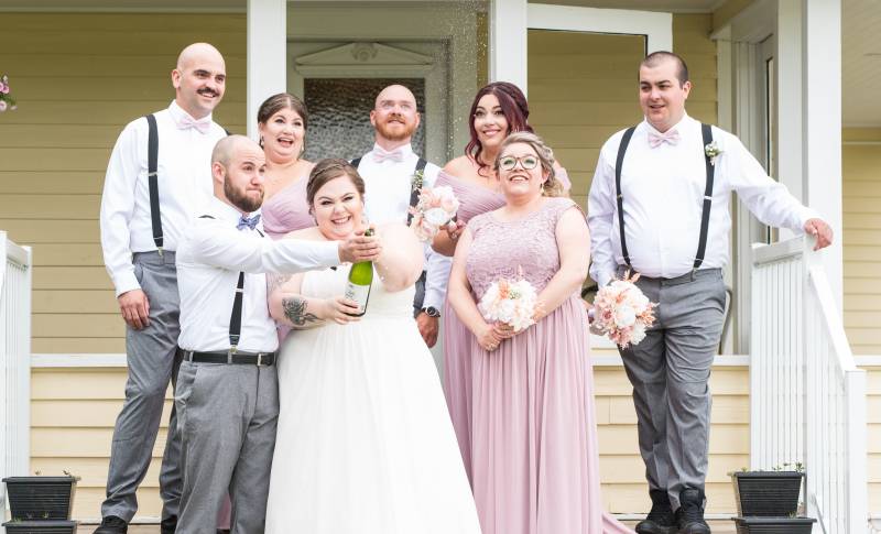 Bride and groom pop green bottle while bridesmaids and groomsmen watch behind 