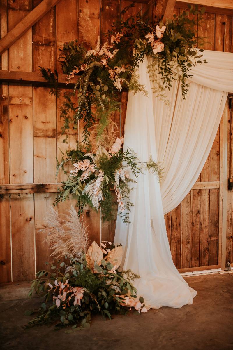 Three blush floral arrangements along edge of white fabric wedding arch 