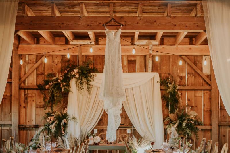 White lace dress hanging on rafters of wooden barn in front of white fabric wedding arch 