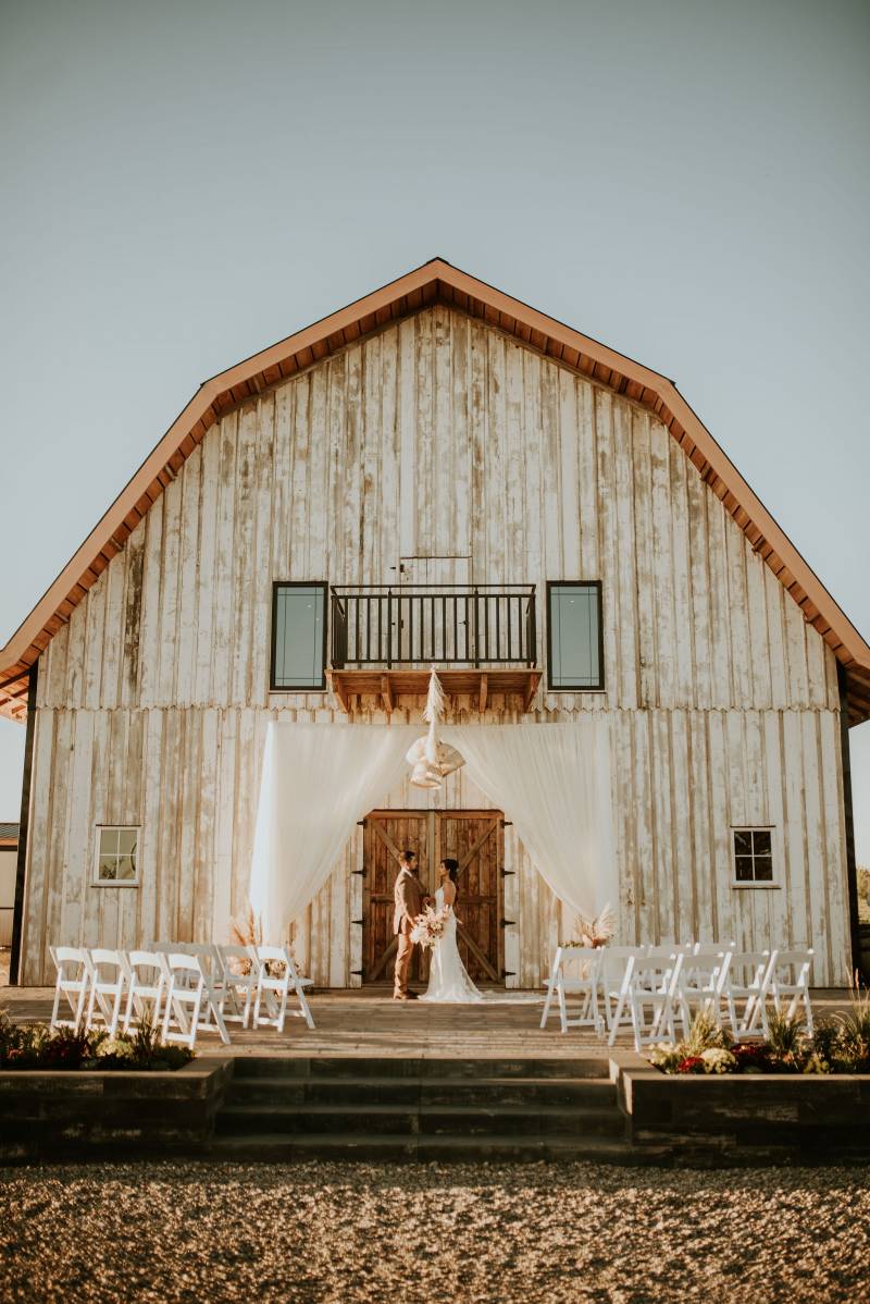 Bride and groom standing underneath white barn and wedding arch with empty seats surrounding them 