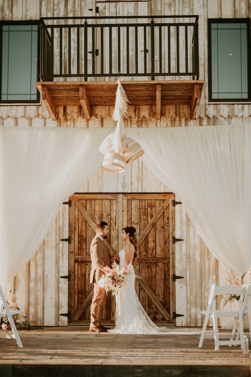 Bride and groom stand underneath white fabric wedding arch and hanging bells in front of wooden door 