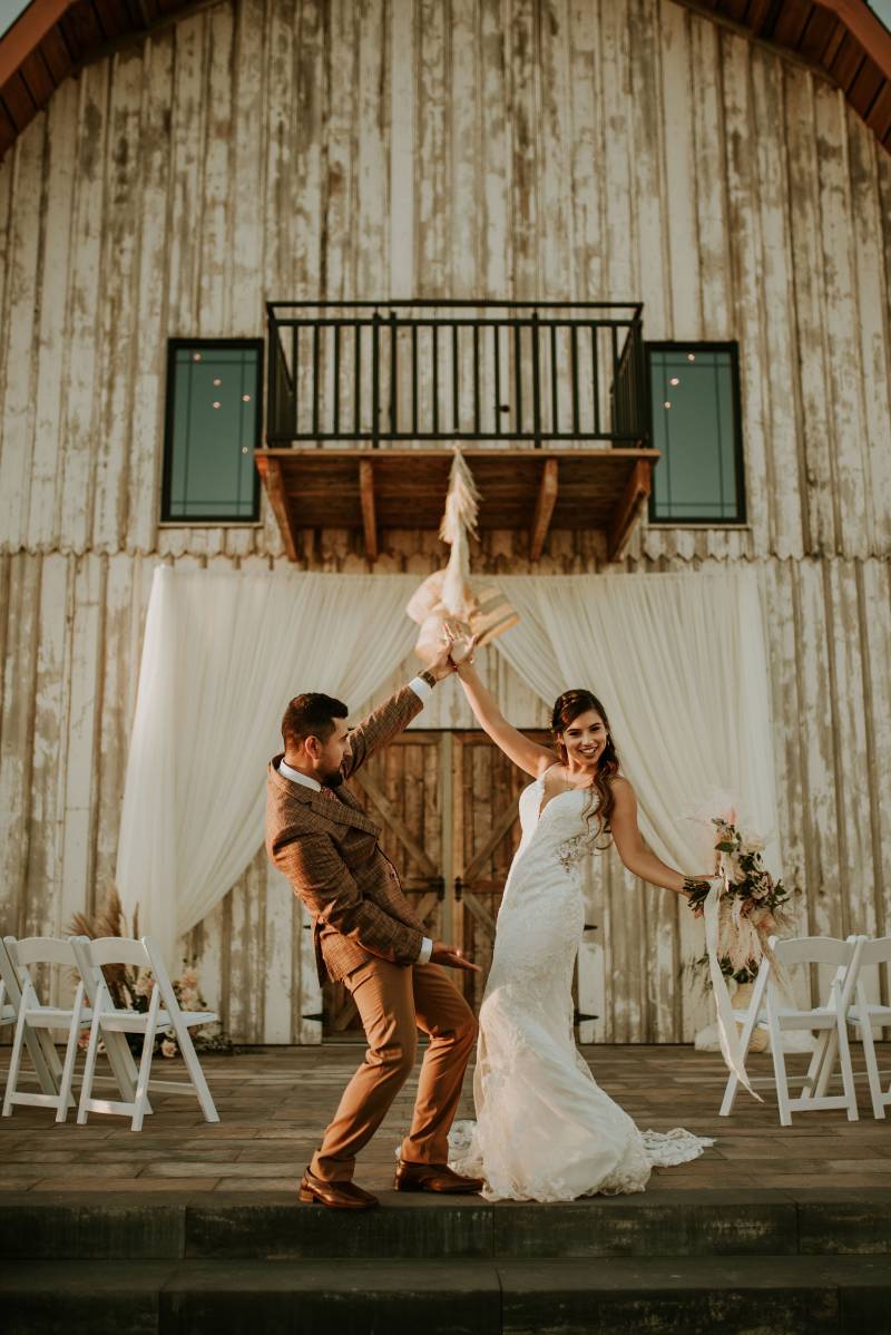 Bride and groom lean back smiling holding hands in front of barn 