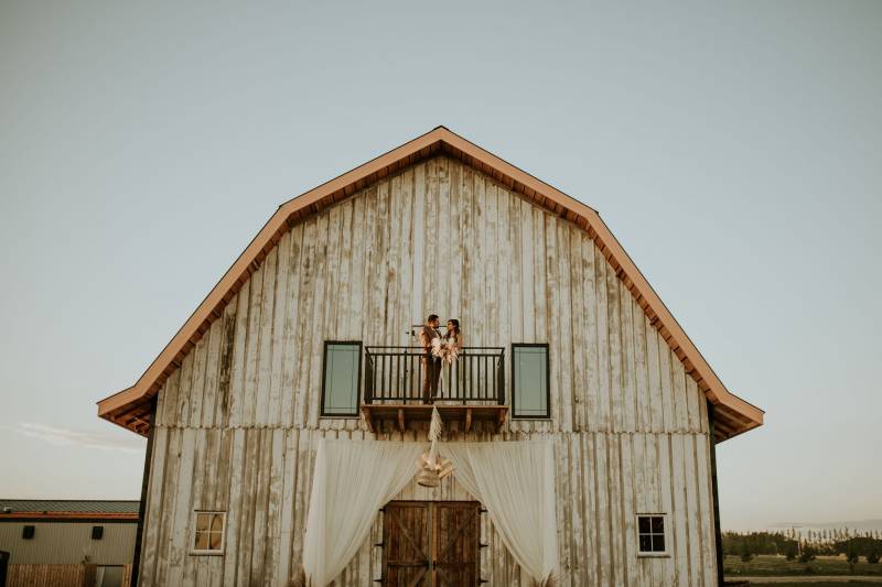 Bride and groom stand on balcony on white barn 