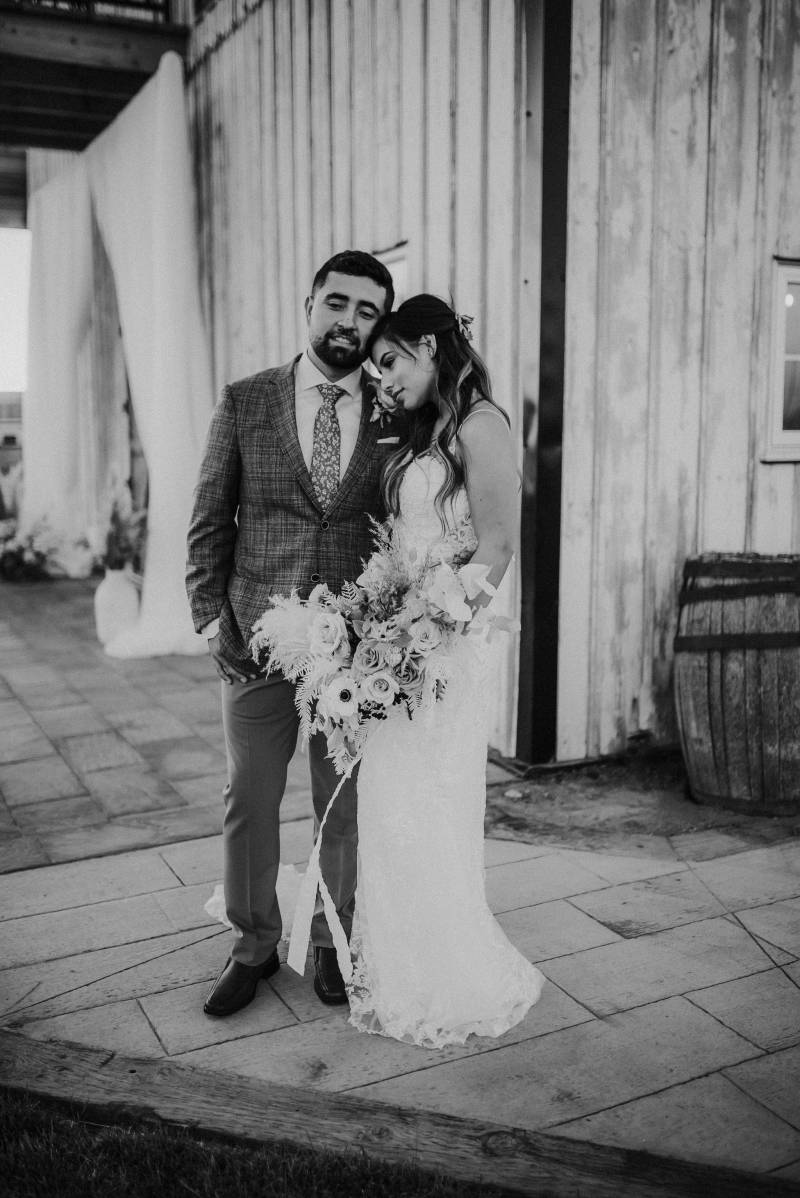 Bride and groom lean against one another in front of barn black and white 