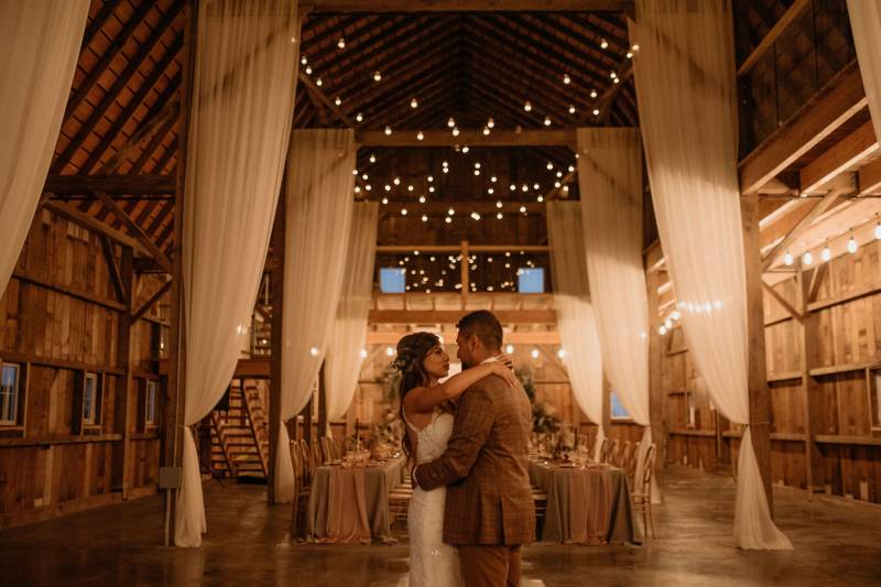 Bride and groom embrace underneath hanging lights and white draping fabric 