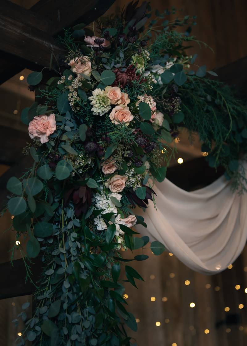 blush pink and burgundy floral arrangement on wedding arch with white fabric