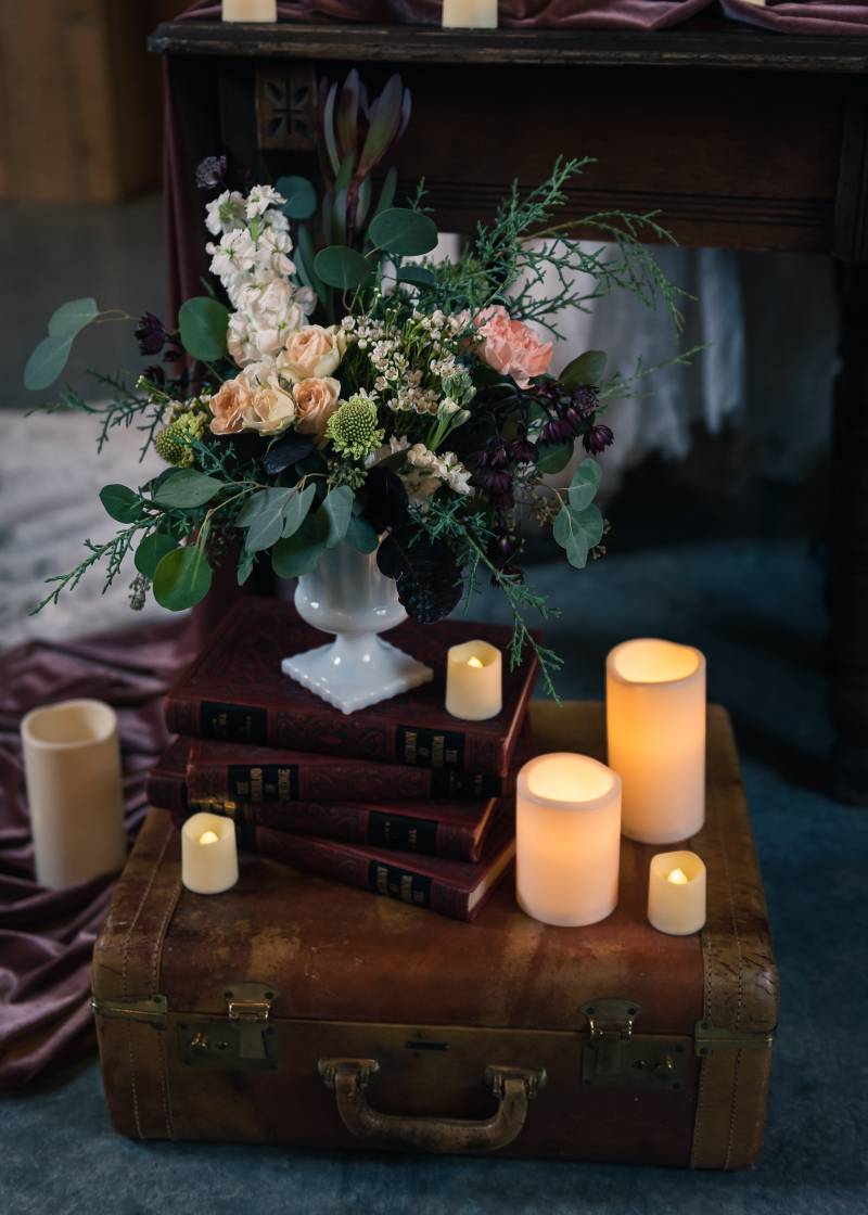 Blush pink and white centerpiece in white vase beside candles on brown suitcase and books 