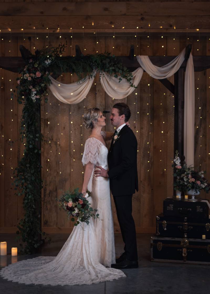 Bride and groom stand facing under dark wood wedding arch and white fabric with fairy lights on wall 