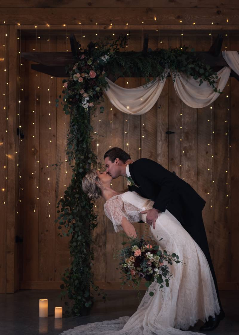 Groom dips bride holding pink and white bouquet under wooden wedding arch and fairy lights on wooden wall