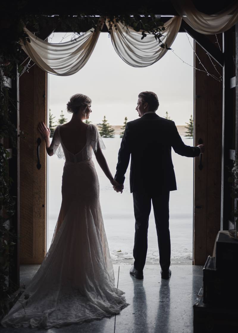 Bride and groom stand holding hands in open doorway under wedding arch with drooping white fabric 