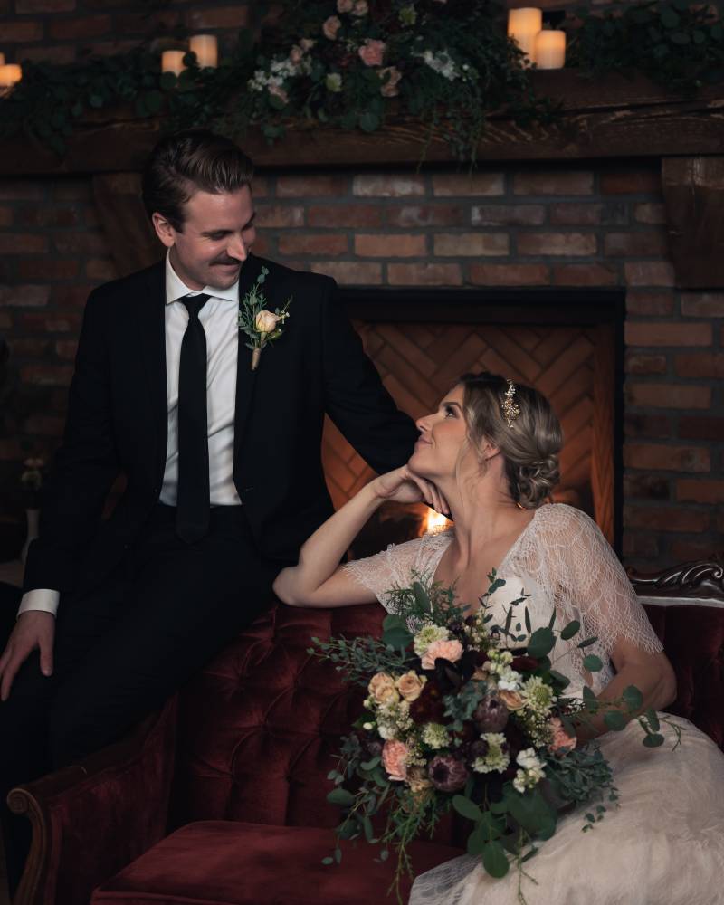 Bride and groom sit looking at one another in front of brick fireplace smiling 