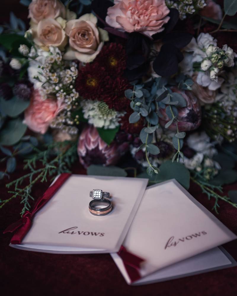 Bride and grooms wedding rings sitting on top of his and her vows in front of pink and burgundy floral arrangement 