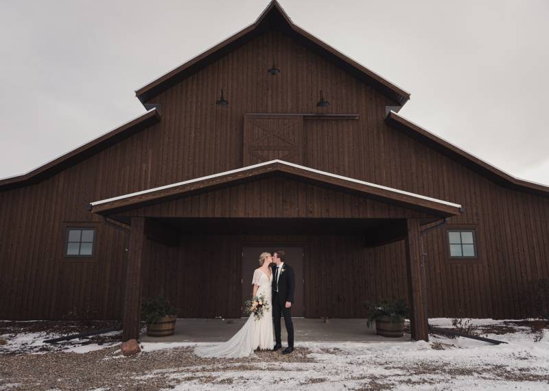 Bride and groom kiss standing in front of large wooden barn on snowy gravel pathway 