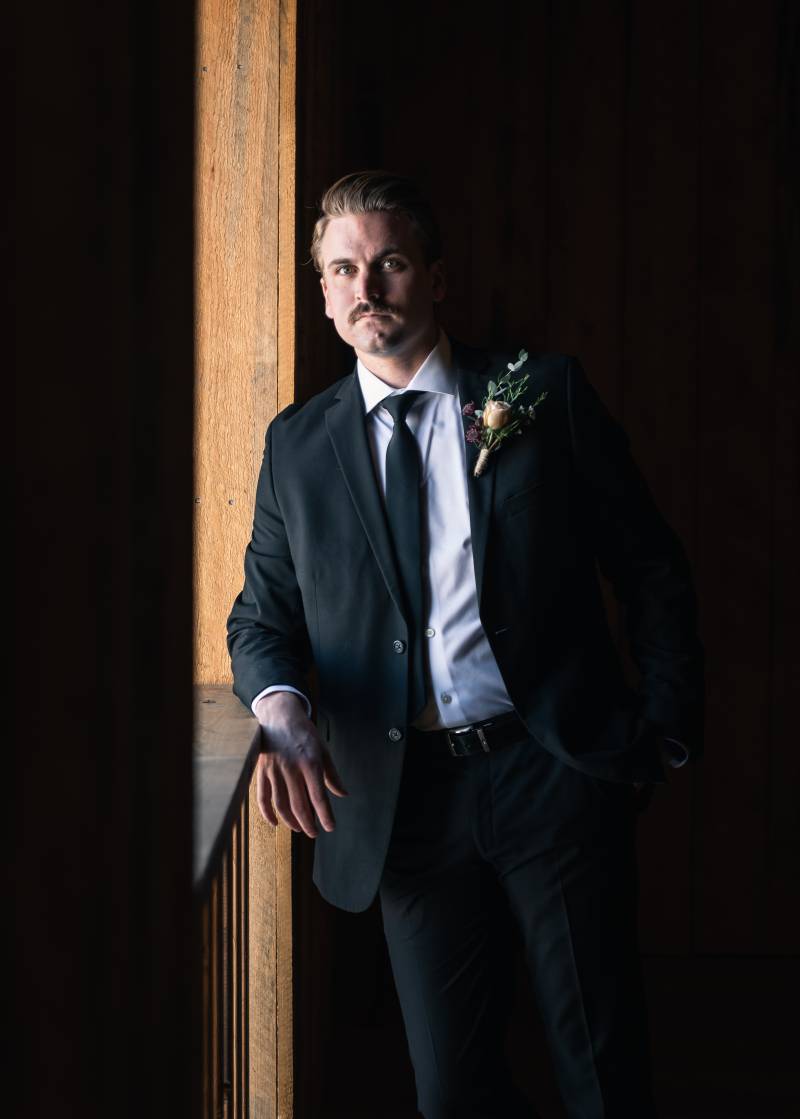 Groom in black suit with green boutonniere leaning against wooden railing 