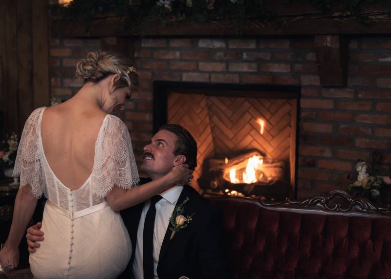 Bride sitting in white open back dress on grooms lap on burgundy sofa in front of brick fireplace