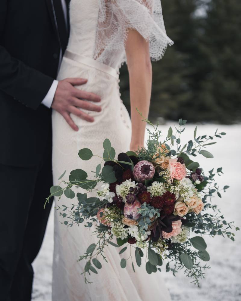 Bride holding bouquet below waist while embraced by groom on snowy field 