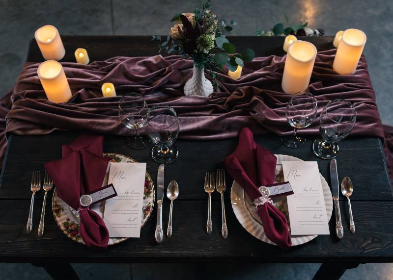 Table setting on small black table with candles and floral centerpiece with silver cutlery and burgundy napkins  