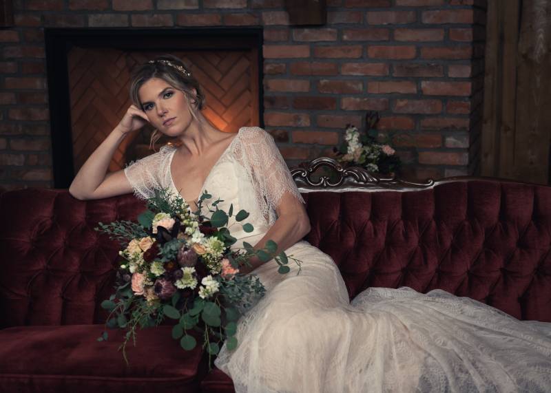 Bride in white lace dress leaning on hand holding pink and white bouquet on burgundy sofa 