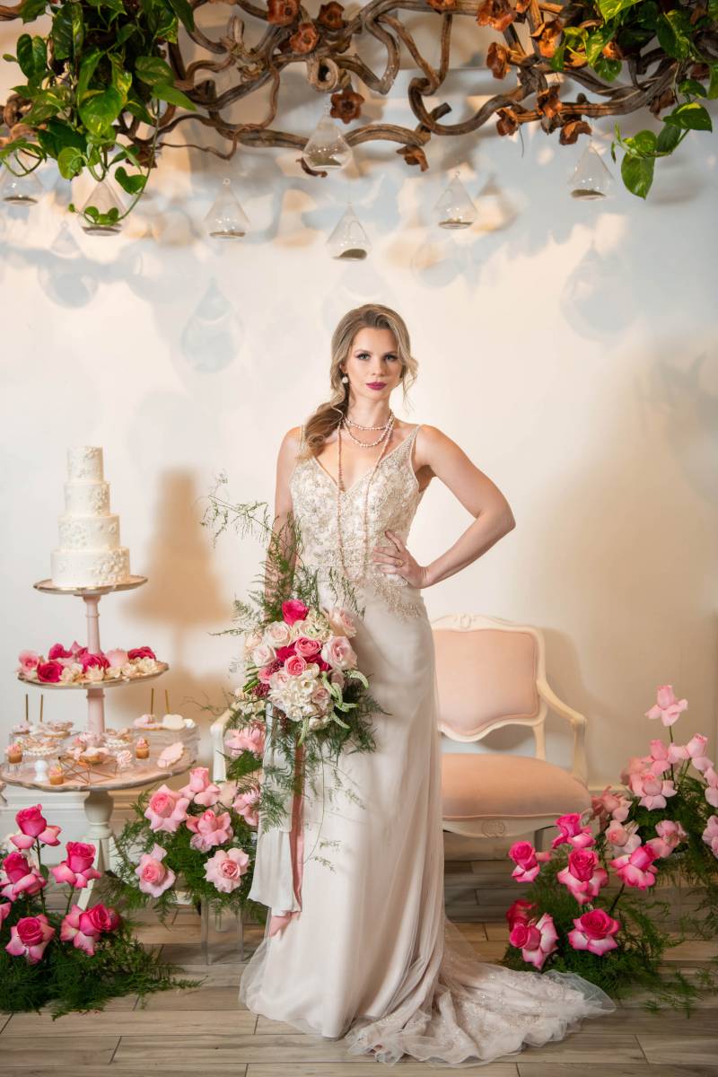 Woman in white dress stands hand on hip holding bouquet in front of pink chair and flowers 