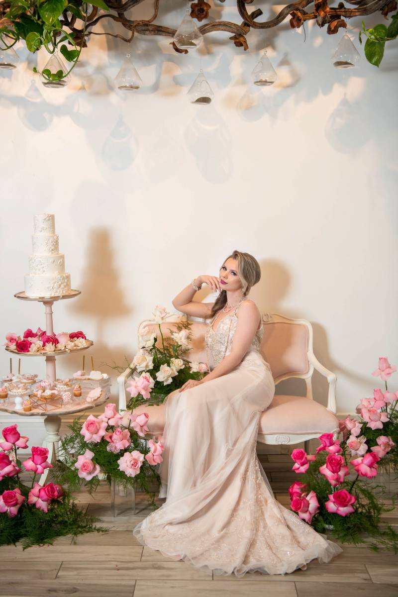 Bride sits in white dress leaning on hand beside white wedding cake arrangement and flowers  