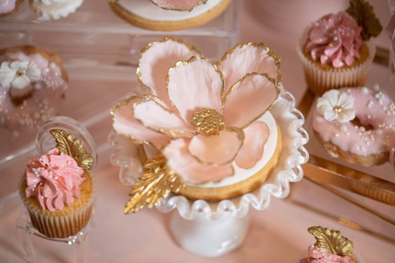 Gold rimmed pink flower in white dish surrounded by pink frosted goods 