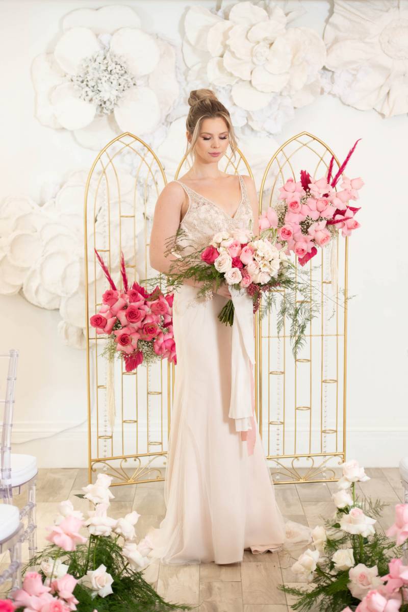 Bride standing holding white bouquet in front of Gold framed arches with pink white and red flowers diagonal across 