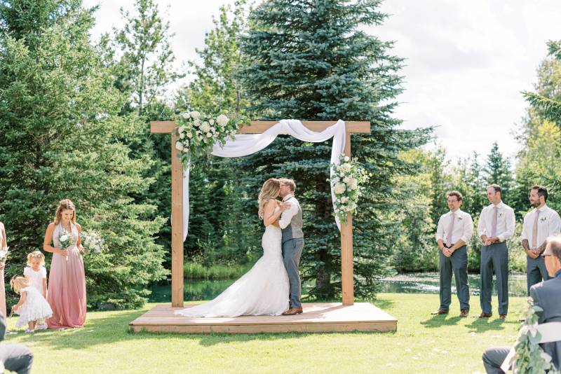 Bride and groom kiss under wedding arch with white fabric while guests watch 